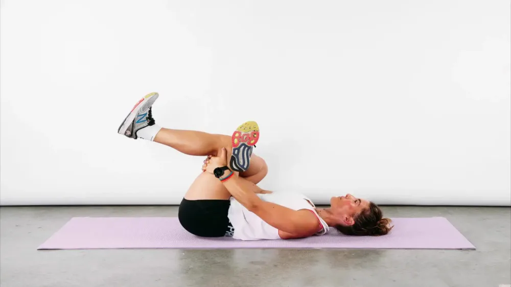 ورزشکار خانم درازکشیده به روی زمین در حال انجام کشش باسن با آوردن یک پا به سمت سینه و قراردادن پای دیگر روی آن